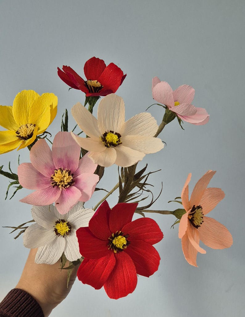 May include: A bouquet of seven paper flowers in various colors, including yellow, pink, red, and white. The flowers are arranged in a hand, with the stems extending out.