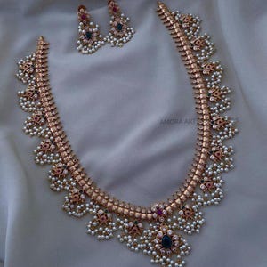May include: A gold necklace with white pearls and colorful gemstones. The necklace has a delicate design with multiple strands of pearls and a central pendant with a large gemstone. The necklace is accompanied by a pair of matching earrings.