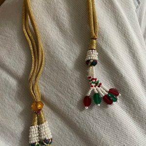 May include: A gold cord necklace with white and red beaded tassels. The tassels have green and red glass beads.
