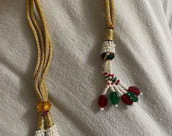 May include: A gold cord necklace with white and red beaded tassels. The tassels have green and red glass beads.