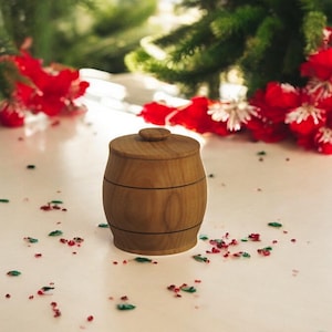 Op de afbeelding: Een klein houten vat met een deksel, dat op een wit oppervlak ligt met rood en groen confetti eromheen.