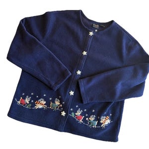 May include: Navy blue fleece cardigan with snowflake buttons and embroidered reindeer design. The reindeer are wearing scarves and skating on a snowy landscape. A cosy, festive sweater for the winter season.