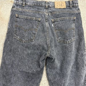 May include: Close-up of the back of acid wash denim jeans. The jeans are a faded black colour with a textured appearance. Features include back pockets, belt loops, and a leather patch. The jeans are a classic style.