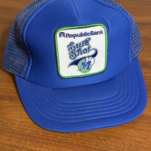 May include: A blue trucker hat with a mesh back and a curved brim. The front of the hat features a patch with the text "RepublicBank Sure Shot M" in white and green. The hat is made of a durable material and is designed to provide shade from the sun.