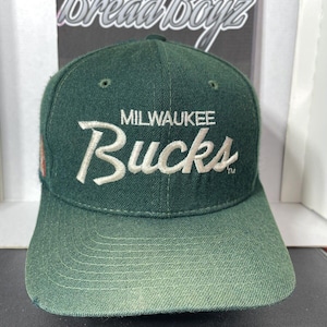 May include: A green baseball cap with the text "MILWAUKEE Bucks" in white script. The cap has a curved brim and a structured crown. The background features the text "BreadBoyz".