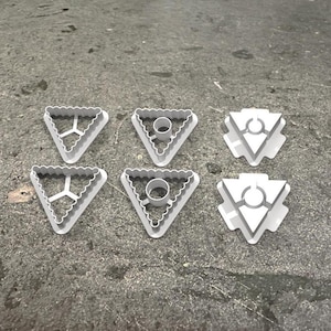 Könnte beinhalten: Sechs weiße, dreieckige Keksausstecher aus Kunststoff mit verschiedenen Designs. Die Designs umfassen einen Kreis mit einem kleineren Kreis im Inneren, eine dreizackige Gabel und einen Kreis mit einer Linie durch ihn.