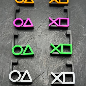 Könnte beinhalten: Vier Sätze von Plastik-Controller-Symbolen, jede in einer anderen Farbe: orange, pink, grün und weiß. Die Symbole sind ein Kreis, ein Dreieck, ein X und ein Quadrat.