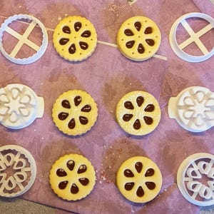 Könnte beinhalten: Ein Set aus neun Ausstechern und sechs Keksen mit Blumenmuster. Die Ausstecher sind aus weißem Kunststoff und die Kekse sind gelb mit brauner Füllung.