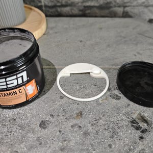 Könnte beinhalten: Geöffnete schwarze Dose ESN Vitamin C-Pulver mit weißem Deckel und separatem schwarzem Deckel. Die Dose hat orangefarbenen und weißen Text. Die Dose ist geöffnet und gibt das Pulver frei. Das Produkt liegt auf einer grauen Oberfläche.
