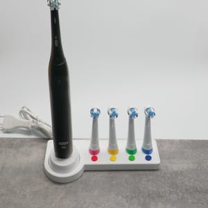 Könnte beinhalten: Eine schwarze elektrische Zahnbürste mit weißer Ladestation und vier Ersatzbürstenköpfen. Die Bürstenköpfe haben farbige Ringe: rosa, gelb, grün und blau. Die Zahnbürste ist mit "Oral B IO" beschriftet.