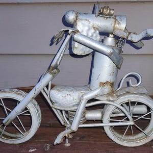 May include: A handcrafted metal sculpture of a motorcycle. The sculpture is made of silver-colored metal with a distressed finish, giving it a vintage look. The motorcycle has two wheels, a seat, handlebars, and a detailed engine.