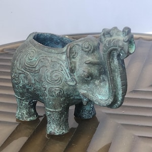 May include: A bronze-colored elephant-shaped container with intricate scrollwork and a weathered patina. The container has a hollowed-out top and is resting on a reflective surface. The elephant has detailed features, including a trunk and tusks.