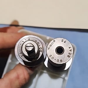 May include: Two silver metal components with circular tops. One has the text "30-06" and "SEAT". The other has the text "30-06" and "L". Both have a screw in the center. The components are held in a clear plastic tray.