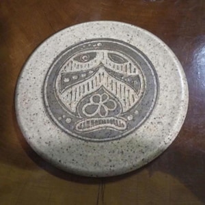 May include: A round ceramic coaster with a speckled off-white finish. The center features a circular gray inlay decorated with an etched, stylized face design that includes a flower motif at the bottom and geometric line patterns throughout.