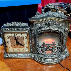 May include: A vintage United brand mantel clock featuring a metallic silver-toned finish with ornate scrollwork. The square clock face has black numerals and hands, while the adjacent section displays a simulated fireplace with glowing orange light.