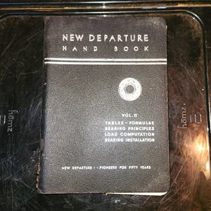 May include: A black, textured hardcover book titled New Departure Hand Book Vol II. The cover features white text detailing tables, formulae, bearing principles, load computation, and bearing installation, with a circular bearing graphic.