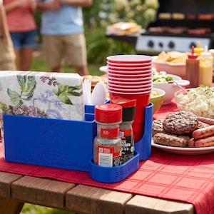 Puede incluir: Un organizador de plástico azul con compartimentos para servilletas, cubiertos y condimentos. El frente presenta el texto en relieve "NAPKINS", "CookoutCaddy" y "10GrationsetRishop", con un icono de mesa de picnic. Un juego de salero y pimentero se encuentra en la bandeja frontal.