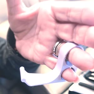 May include: A white, 3D-printed finger hook tool. The tool is designed to assist with opening doors or pressing buttons without direct contact. The tool is held by a finger, and the hook extends outward.