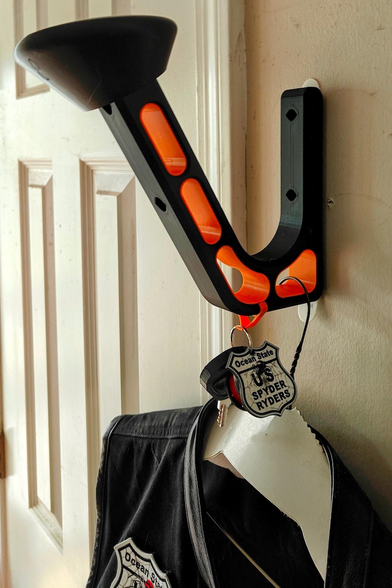 May include: Black and orange wall-mounted coat rack with a curved design. The rack has a key holder and is holding a black jacket. The key chain has the text "Ocean State US SPYDER RYDERS".