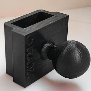 Puede incluir: Un objeto negro, rectangular, impreso en 3D con un pomo esférico adjunto. El objeto tiene una abertura rectangular en la parte superior y la palabra "PRINT" es visible en el lateral. Es probable que sea un molde o un componente para un proyecto más grande.