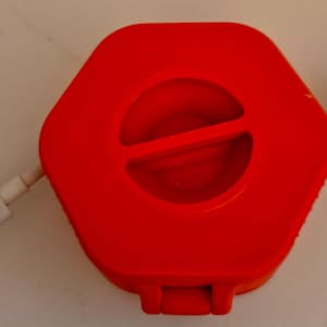 May include: A red, hexagonal cable organizer with a central circular opening and a white USB cable extending from each side. The organizer has a small latch on the bottom. The cable is white.