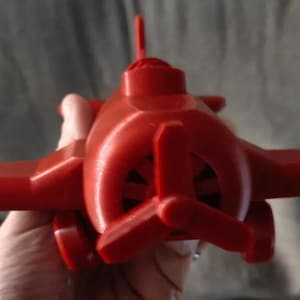 May include: A red plastic toy airplane with a propeller. The airplane has a simple design with a rounded body and wings.