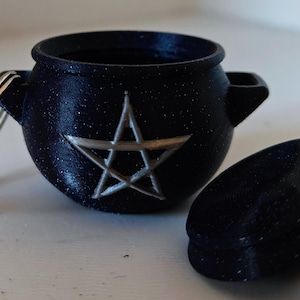 May include: A black cauldron keychain with a silver pentagram design. The cauldron has a lid and a silver key ring.