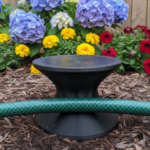 Puede incluir: Un carrete de manguera negro con una manguera verde, colocado en un jardín con flores coloridas. El carrete tiene una base ancha y una parte superior ensanchada, diseñada para sujetar una manguera de jardín. La manguera es verde con un patrón de diamantes.