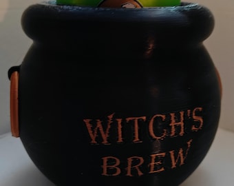 May include: Black cauldron-shaped beverage holder with the text "Witch's Brew" in copper-colored lettering. A green can with the text "Refreshingly Natural" is sitting inside the cauldron.