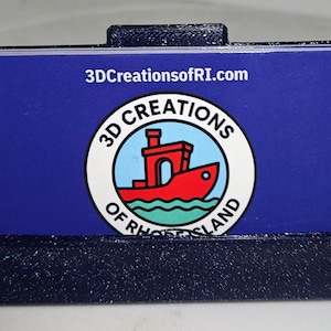 May include: A dark blue 3D-printed business card holder containing a blue business card. The card features the text 3DCreationsofRI.com and a circular logo depicting a red boat on light blue waves with the text 3D CREATIONS OF RHODE ISLAND.