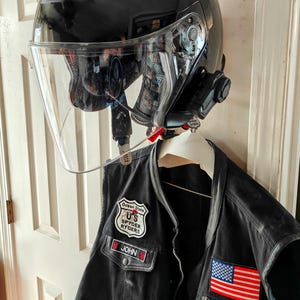 May include: A black motorcycle helmet with a clear visor and a communication system attached, hanging above a black leather vest. The vest features patches including the American flag and "Rhode Island."