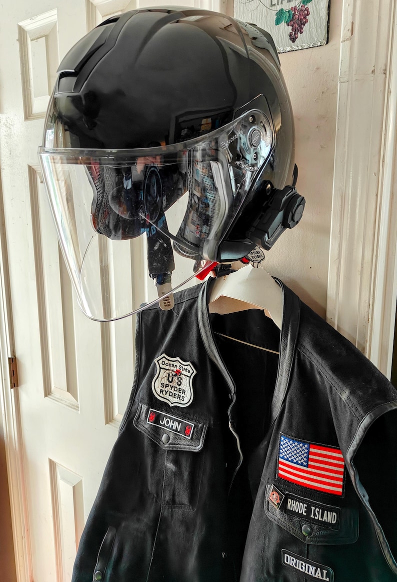 May include: A black motorcycle helmet with a clear visor and a communication system attached, hanging above a black leather vest. The vest features patches including the American flag and "Rhode Island."