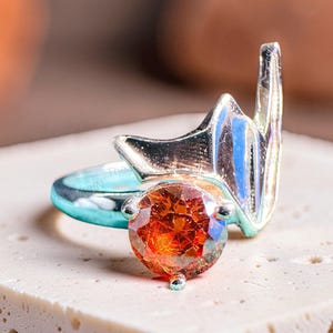 May include: A silver-toned ring featuring a round, faceted orange gemstone set in a prong setting. The band extends into a stylized, pointed wing-like structure that rises vertically from the side of the central stone, creating a distinct silhouette.