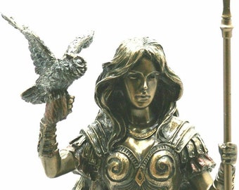 Può includere: Una statua di bronzo di una guerriera con un mantello rosso fluttuante, che tiene una lancia in una mano e un gufo nell'altra. La guerriera indossa un'armatura con dettagli intricati e una corona dorata.