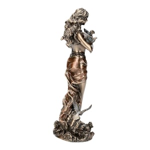 May include: A bronze sculpture of a woman holding a bird in her arms. The woman is standing on a wave-like base. The sculpture is painted in shades of brown and gold.