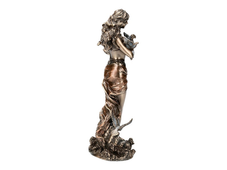 May include: A bronze sculpture of a woman holding a bird in her arms. The woman is standing on a wave-like base. The sculpture is painted in shades of brown and gold.
