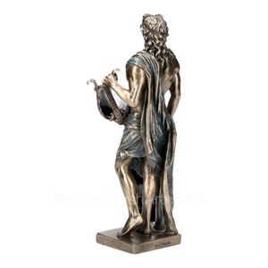 Puede incluir: Una estatua de tono bronce de una figura con cabello largo y rizado, de pie sobre una base rectangular. La figura est&aacute; envuelta en una prenda de tela con detalles azules y sostiene una lira en una mano. El texto BronzeFinishArtworks es visible.