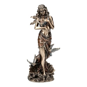 May include: A bronze sculpture of a woman standing on a wave with birds flying around her. The woman is wearing a flowing robe and has long flowing hair. The sculpture is detailed and intricate.
