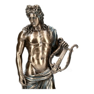 Puede incluir: Una estatua con acabado en bronce de una figura con cabello largo y ondulado, que lleva tela drapeada alrededor de la cintura y el hombro. La figura sostiene una lira en la mano izquierda, con intrincadas tallas decorativas en el marco.