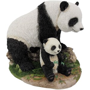May include: A figurine of a panda bear and cub. The adult panda is mostly white with black markings and is seated. The cub is in front, also with black and white markings. The base is a rock-like structure with green foliage.