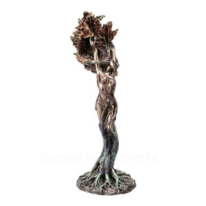 May include: A bronze-toned sculpture depicting a figure merging with a tree. The base shows exposed roots, transitioning into a trunk-like torso with branches and leaves extending upward from the head and arms, finished in metallic brown and green.