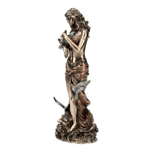May include: Bronze sculpture of a woman standing on a wave with birds flying around her. The woman is holding a bird in her hand and has a flowing robe.
