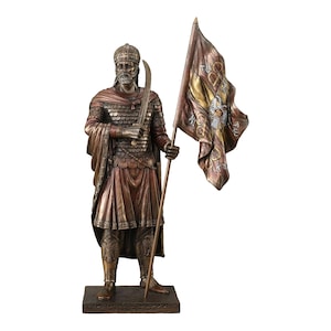 May include: A bronze-toned statue of a warrior wearing scale armor, a helmet, and a cloak, holding a curved sword in one hand and a flag with gold and brown fabric in the other. The base features the text KONSTANTINOS XI PALAIOLOGOS 9.2.1404-29.5.1453.