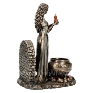 Puede incluir: Una estatua de bronce de una mujer sosteniendo una llama, de pie frente a un arco de piedra y un caldero con un dise&ntilde;o de nudo celta. La estatua est&aacute; sobre una base con fuego debajo del caldero.