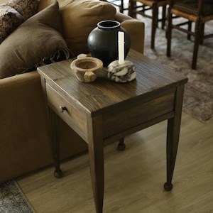 May include: A dark brown wooden end table with a single drawer and turned legs. The table is decorated with a black ceramic vase, a white candle, a small wooden bowl, and a white marble object.