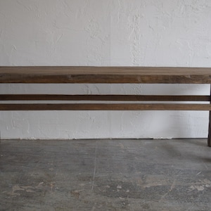 May include: A rustic wooden bench with a simple design. The bench has a rectangular top and four legs, with a single stretcher running across the bottom. The wood is a dark brown color and has a weathered finish.