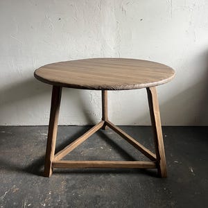 May include: A round wooden table with a natural brown finish and a textured edge. The table features three straight legs connected by a triangular support frame near the base, providing a stable structure for the circular tabletop surface.
