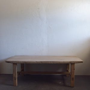 May include: A light-colored, rectangular wooden coffee table with four sturdy legs and a lower support beam. The table has a natural wood grain finish and is set against a white wall.