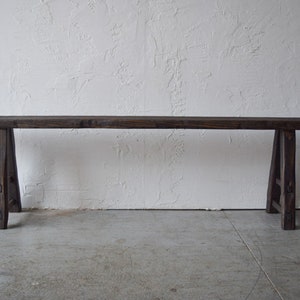 May include: A rustic, dark brown wooden bench with a rectangular top and angled legs. The bench is made of weathered wood, showcasing natural grain and knots. The design is simple and sturdy, suitable for indoor or outdoor use.