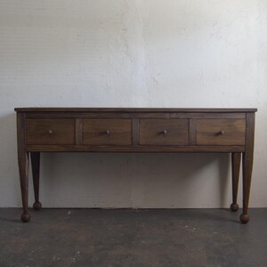 May include: A wooden console table with four drawers and a dark brown finish. The table has slender, tapered legs with ball feet. The drawers have small, round knobs. The table is set against a white wall.